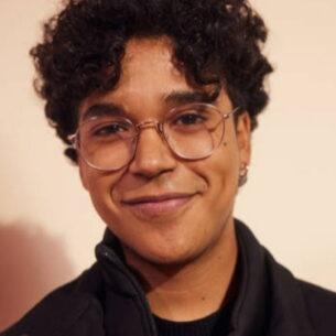 A person with curly dark hair and round glasses smiling.