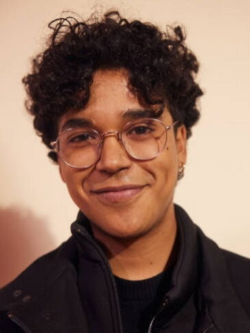 A person with curly dark hair and round glasses smiling.